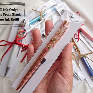 Peut inclure: Un stylo en or rose avec des fleurs s&eacute;ch&eacute;es ench&acirc;ss&eacute;es dans de la r&eacute;sine transparente. Le stylo est emball&eacute; dans une enveloppe blanche avec le texte "Recharge d'encre uniquement ! Choisissez entre l'encre noire ou bleue".