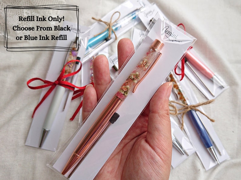 Peut inclure: Un stylo en or rose avec des fleurs s&eacute;ch&eacute;es ench&acirc;ss&eacute;es dans de la r&eacute;sine transparente. Le stylo est emball&eacute; dans une enveloppe blanche avec le texte "Recharge d'encre uniquement ! Choisissez entre l'encre noire ou bleue".