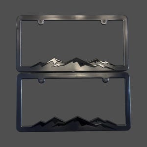 May include: Two license plate frames with a mountain range design. The top frame is silver, and the bottom frame is black. Both frames have a rectangular shape with rounded corners and mounting holes.