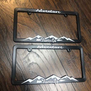 May include: Two black metal license plate frames with white text and a mountain range design. The top frame says "Adventure Awaits" and the bottom frame says "Adventure Awaits".