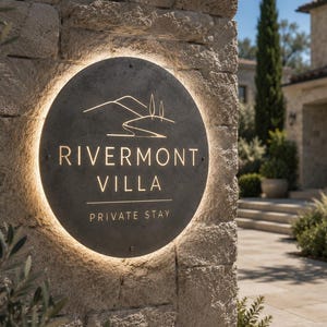 Puede incluir: Un letrero circular de piedra gris oscuro montado en una pared de piedra texturizada, con el texto RIVERMONT VILLA y PRIVATE STAY en letras doradas, junto a un dibujo lineal de colinas y árboles, iluminado por una luz de fondo cálida.