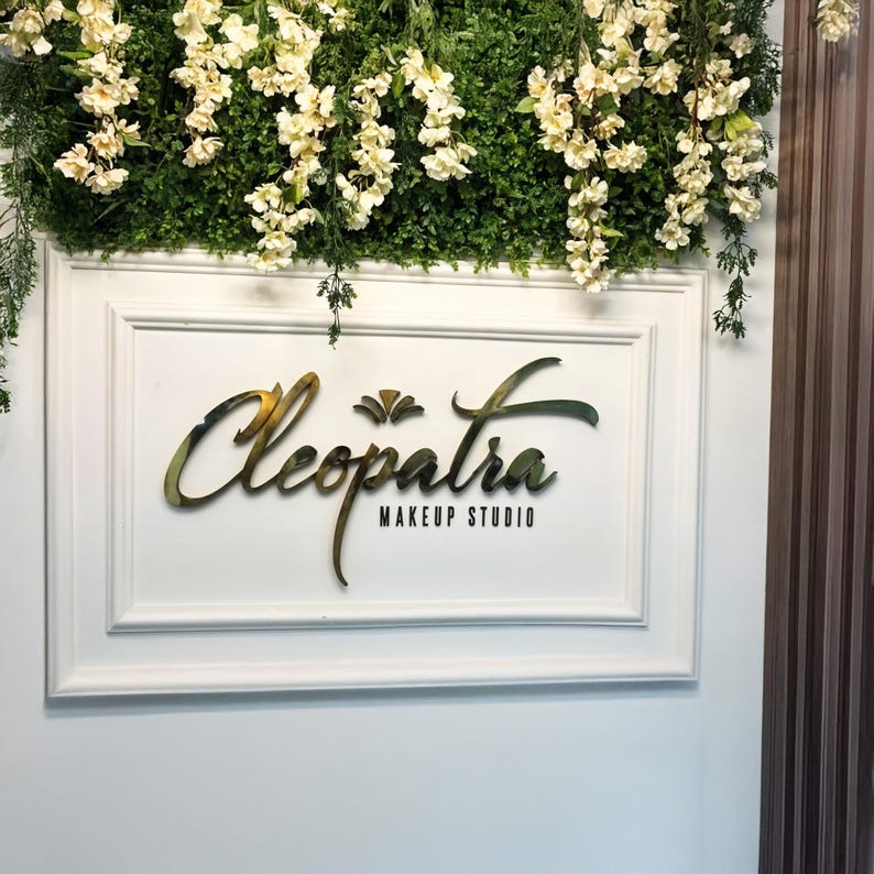 Cleopatra Makeup Studio sign in gold gradient letters framed with white molding and floral greenery – chic makeup artist wall decor