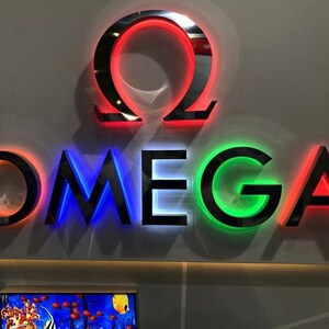 May include: A black and silver sign with the word "OMEGA" in white letters with red, blue, and green lights behind each letter. The OMEGA logo is a red circle with a black omega symbol inside.