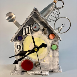 May include: A house-shaped clock constructed from gray textured material, featuring a central yellow dial with black hands. It is decorated with a metal spoon, a circular thermometer, wire coils, and colorful circular accents in red, orange, green, and blue.