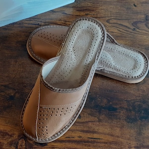 Op de afbeelding: Een paar bruine leren slippers met een geperforeerd ontwerp. De slippers hebben een zacht, gevoerd voetbed en een comfortabele pasvorm.