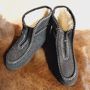May include: A pair of gray felted wool slippers with white stitching and a zipper closure. The interior is lined with white shearling. The slippers are designed for indoor use and provide warmth and comfort.