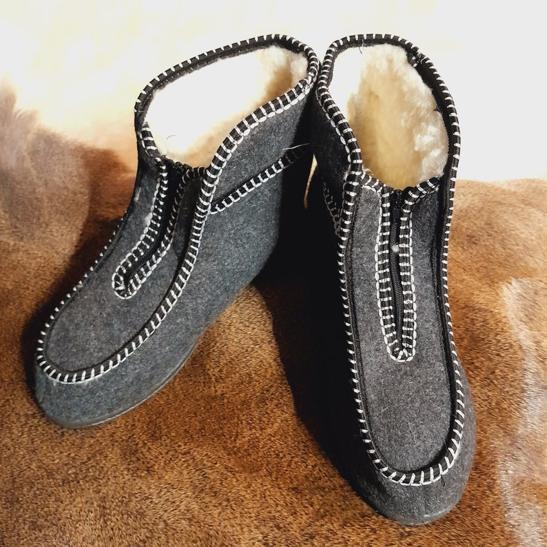 May include: A pair of gray felted wool slippers with white stitching and a zipper closure. The interior is lined with white shearling. The slippers are designed for indoor use and provide warmth and comfort.