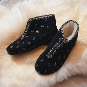 Könnte beinhalten: Ein Paar schwarze Hausschuhe mit floralem Muster und Reißverschluss. Die Stiefel haben eine weiß-graue Bordüre und ein weiches, weißes Innenfutter. Die Stiefel sind für den Innenbereich konzipiert.