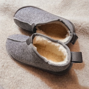 May include: A pair of gray felt slippers with a soft, cream-colored interior. The slippers have a leather trim and a loop on the heel. The slippers are designed for indoor use and provide warmth and comfort.