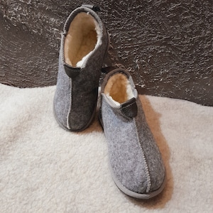 May include: A pair of gray felt slippers with a white fleece lining. The slippers have a rounded toe and a closed back.
