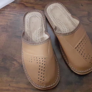 Op de afbeelding: Een paar bruine leren slippers met een geperforeerd ontwerp. De slippers hebben een gesloten neus en een open achterkant om in te schuiven.