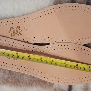 May include: Two brown leather insoles with a perforated pattern. The insoles are measured with a yellow tape measure, showing a length of 10 inches. The insoles are labeled "BONTES UBER" and "41".