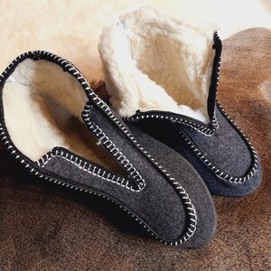 May include: A pair of gray felt slippers with white stitching and a white, fluffy interior. The slippers have a zipper closure and are designed for indoor use. The slippers are shown on a brown surface.