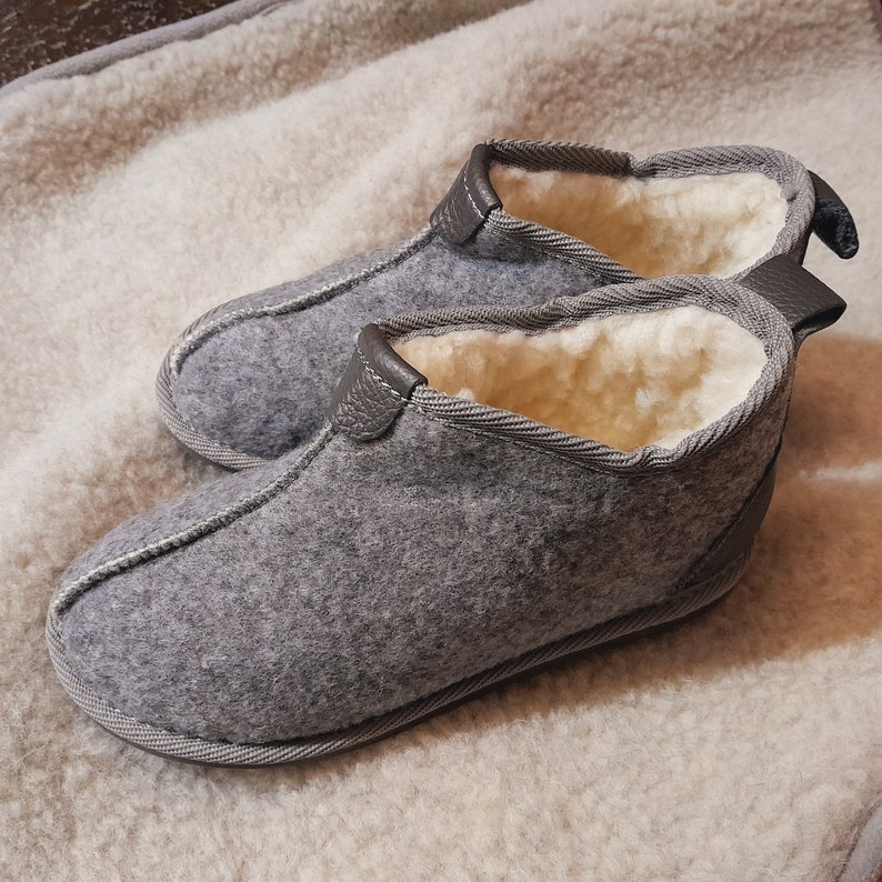 May include: A pair of gray felt slippers with a soft, cream-colored interior. The slippers have a leather trim and a stitched design around the edges. The slippers are designed for indoor use.