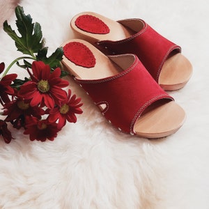 May include: A pair of red open-toe sandals with wooden soles and white stitching. The sandals have a red suede-like upper and a decorative red patch on the insole. A bouquet of red flowers with green leaves is next to the sandals.
