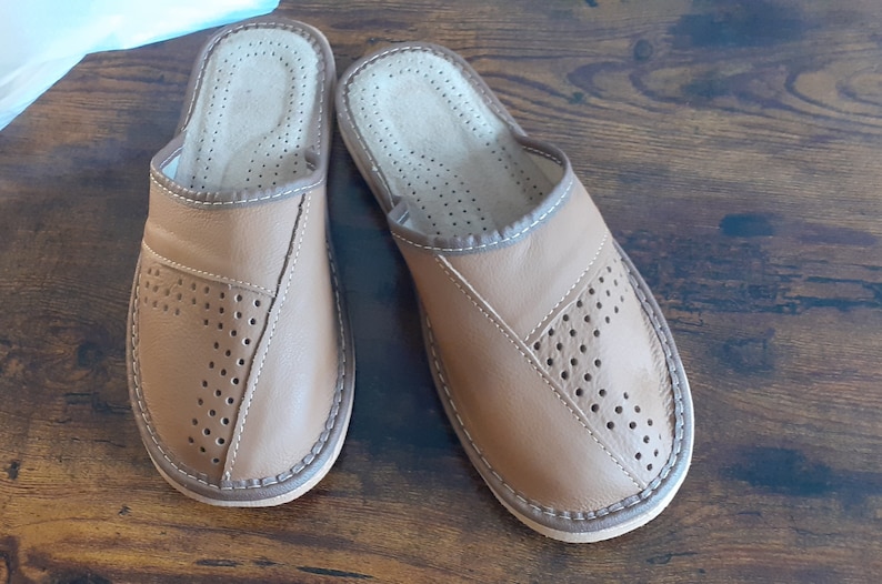 Op de afbeelding: Een paar bruine leren slippers met een geperforeerd ontwerp. De slippers hebben een witte stikdetail rond de randen.
