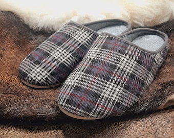 May include: A pair of plaid pattern slippers in gray, black, red, and white. The slippers have a gray trim and a light-colored sole. The background is a mix of brown and white textures.