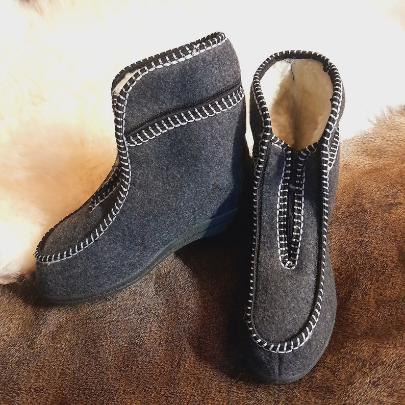 May include: A pair of dark gray felt boots with white stitching and a zipper closure. The boots have a soft, light-colored interior and a black sole. The boots are designed for indoor use.
