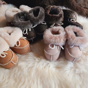 Könnte beinhalten: Ein Set mit sechs Paar Babyschuhe aus Schaffell in Braun, Beige und Rosa. Die Schuhe haben ein weiches, flauschiges Futter und eine Ledersohle.