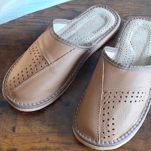 Op de afbeelding: Een paar bruine leren slippers met een geperforeerd ontwerp. De slippers hebben een gesloten neus en een open achterkant. De slippers liggen op een houten oppervlak.
