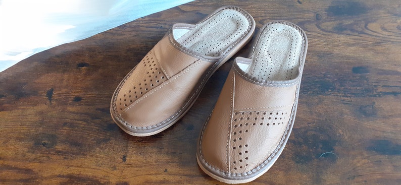 Op de afbeelding: Een paar bruine leren slippers met een geperforeerd ontwerp. De slippers hebben een gesloten neus en een open achterkant. De slippers liggen op een houten oppervlak.