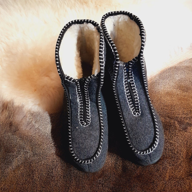 May include: A pair of dark gray felted wool slippers with white stitching and a white fleece lining. The slippers have a zipper closure and are designed to keep feet warm. The slippers are shown on a brown and beige surface.
