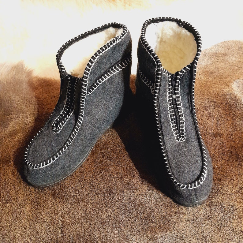 May include: A pair of gray felt boots with white stitching and a zipper closure. The boots have a soft, white interior lining and are designed for warmth and comfort. The boots are shown on a brown surface.