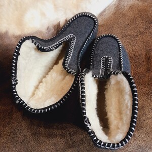 May include: A pair of gray felt slippers with white shearling lining. The slippers have a black trim with white stitching. The slippers are designed for indoor use and provide warmth and comfort. The slippers are shown on a brown surface.