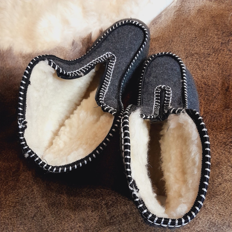 May include: A pair of gray felt slippers with white shearling lining. The slippers have a black trim with white stitching. The slippers are designed for indoor use and provide warmth and comfort. The slippers are shown on a brown surface.