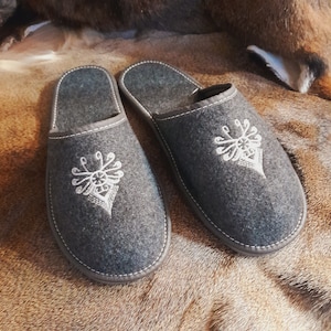 Peut inclure: Une paire de pantoufles en feutre gris avec des motifs brodés blancs. Les pantoufles ont une pointe fermée et un style à enfiler.
