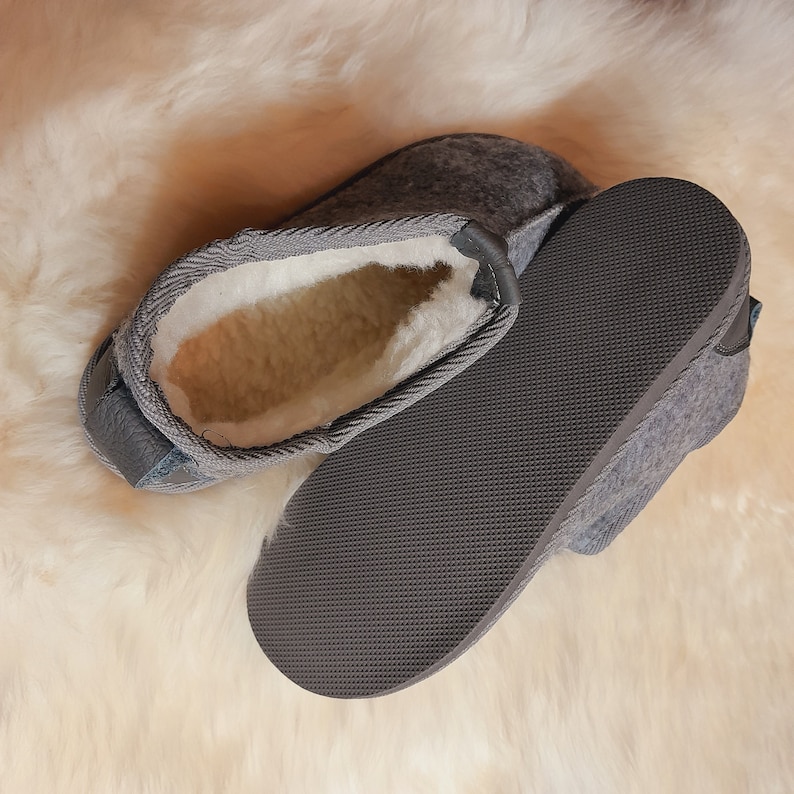 May include: Gray felt slippers with a white, fluffy interior and a dark gray rubber sole. The slippers are open and displayed on a cream-colored, textured surface. The slippers have a leather trim.