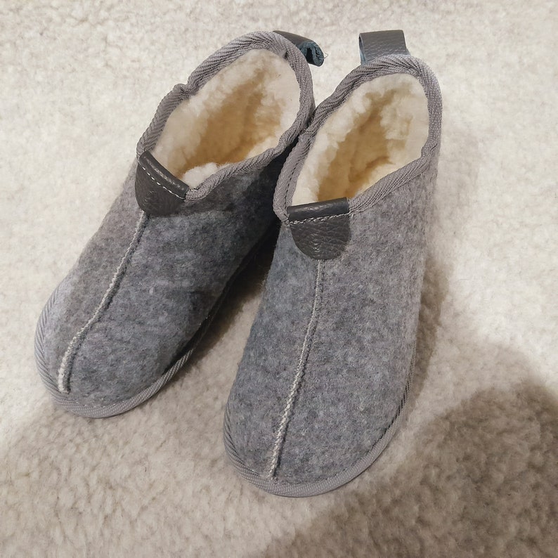 May include: A pair of gray felt slippers with a soft, cream-colored interior lining. The slippers have a rounded toe and a leather accent on the heel. The slippers are designed for indoor use and provide warmth and comfort.