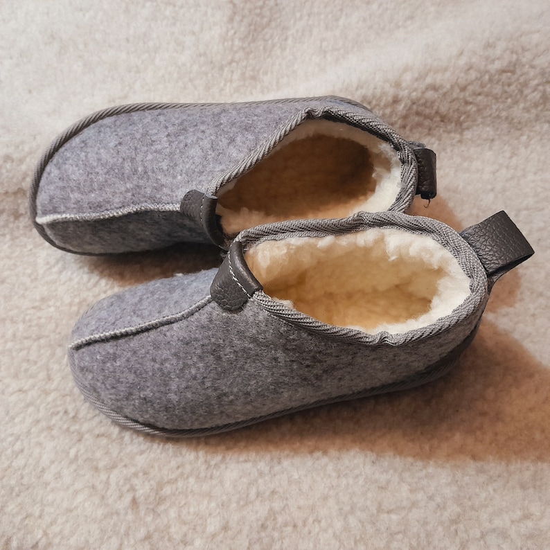 May include: A pair of gray felt slippers with a soft, cream-colored interior. The slippers have a leather accent on the heel and are designed for indoor use. The slippers are made of a soft, warm material.