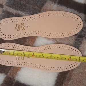 May include: Two beige leather insoles with a pattern of small holes. The insole on the bottom has a yellow measuring tape showing a length of 10 inches. The insole on top has the number 37 printed on it.