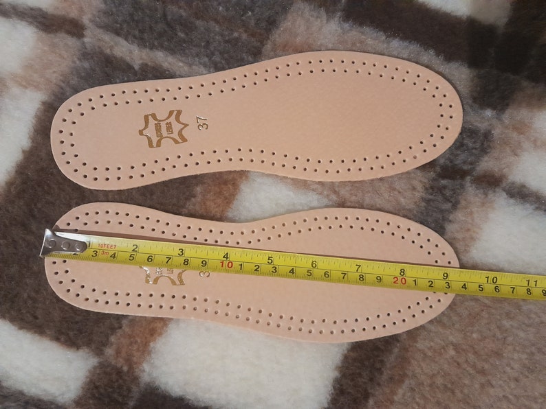 May include: Two beige leather insoles with a pattern of small holes. The insole on the bottom has a yellow measuring tape showing a length of 10 inches. The insole on top has the number 37 printed on it.