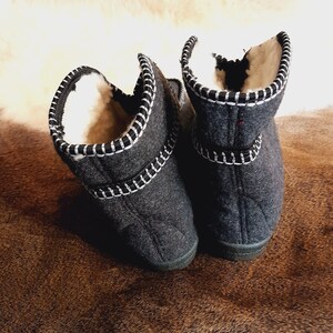 May include: A pair of dark gray felted wool slippers with white stitching and a black trim. The interior is lined with a cream-colored, fluffy material. The slippers are designed to be worn indoors and provide warmth and comfort.