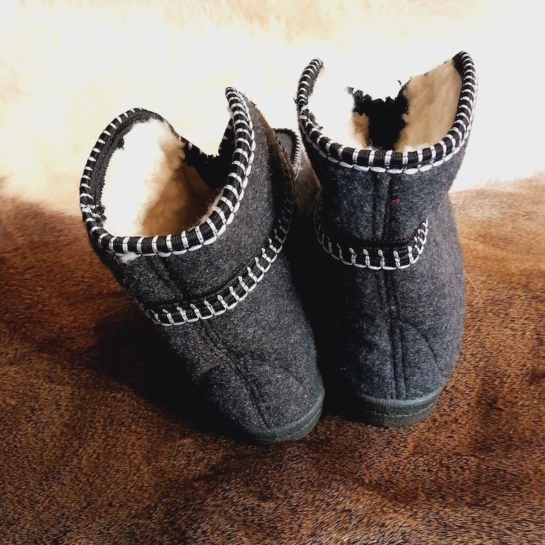 May include: A pair of dark gray felted wool slippers with white stitching and a black trim. The interior is lined with a cream-colored, fluffy material. The slippers are designed to be worn indoors and provide warmth and comfort.