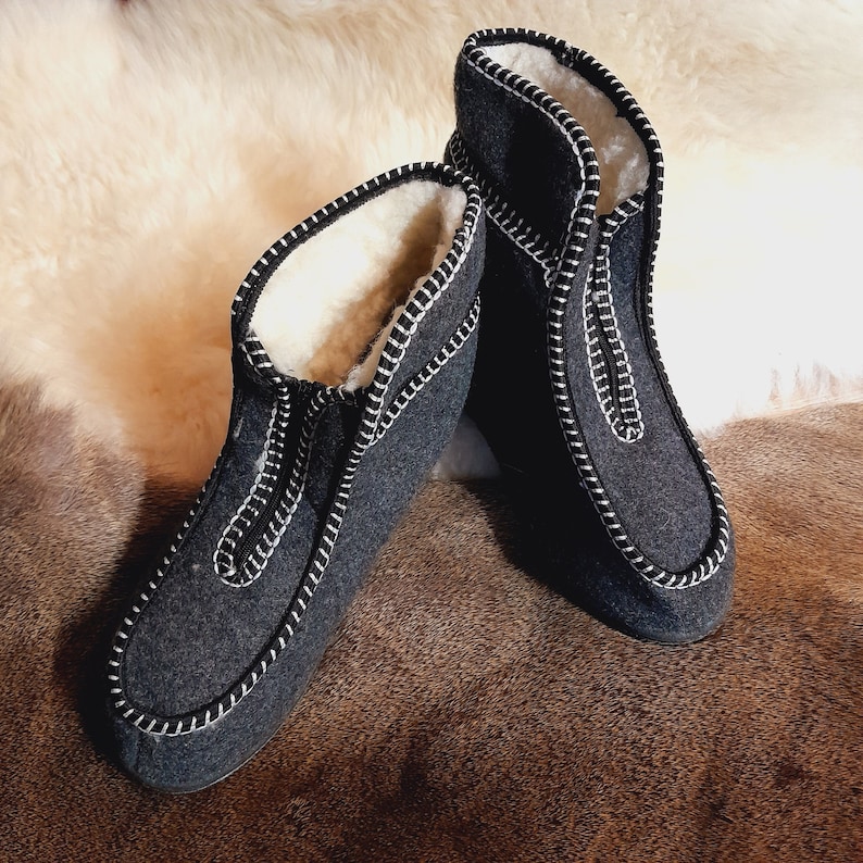 May include: A pair of dark gray felted wool slippers with white stitching and a white fleece lining. The slippers have a zip-up front and are designed for indoor use. The background features a light-colored fur.
