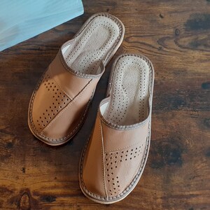 Op de afbeelding: Een paar bruine leren slippers met een geperforeerd ontwerp. De slippers hebben een gesloten neus en een open achterkant. De slippers liggen op een houten oppervlak.