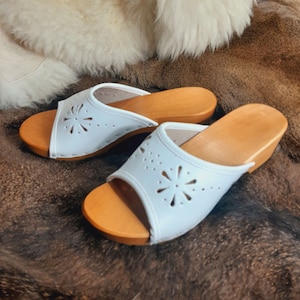 Könnte beinhalten: Ein Paar weiße Lederclogs mit einem floralen Ausschnittmuster auf den Riemen. Die Clogs haben eine Holzsohle und einen kleinen Absatz. Die Schuhe werden auf einer braunen, strukturierten Oberfläche präsentiert.