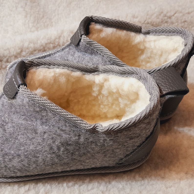 May include: A pair of gray felt slippers with a light-colored, fluffy interior. The slippers have a strap detail and are designed for indoor wear. The felt material is a soft gray, and the interior appears to be made of wool or a similar material.