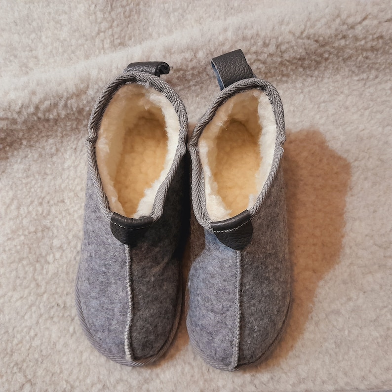 May include: A pair of gray felt slippers with a soft, cream-colored interior. The slippers have a leather pull tab at the back and a stitched seam detail. The slippers are designed for indoor use and provide warmth and comfort.