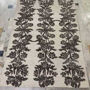 May include: A rectangular area rug featuring a light beige background with three vertical columns of dark brown, raised floral and vine patterns. The design consists of repeating botanical motifs that run the full length of the rug.