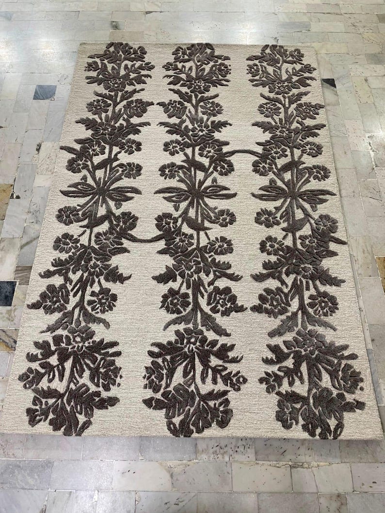 May include: A rectangular area rug featuring a light beige background with three vertical columns of dark brown, raised floral and vine patterns. The design consists of repeating botanical motifs that run the full length of the rug.