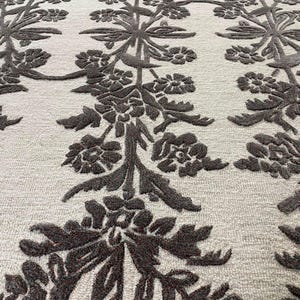 May include: A rectangular area rug featuring a light cream-coloured base with a repeating vertical pattern of dark brown, raised floral and vine motifs. The design consists of symmetrical botanical elements that create a textured, high-contrast surface.
