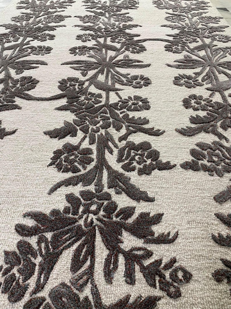 May include: A rectangular area rug featuring a light cream-coloured base with a repeating vertical pattern of dark brown, raised floral and vine motifs. The design consists of symmetrical botanical elements that create a textured, high-contrast surface.