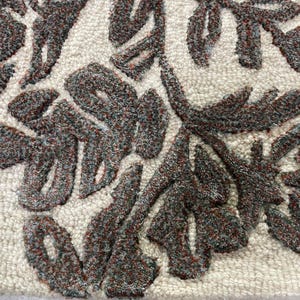 May include: A tufted textile featuring a cream-coloured background with a raised, dark charcoal and rust-coloured floral pattern. The design consists of intricate, textured leaf and petal shapes created through a dense loop pile construction.