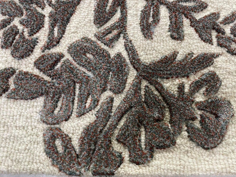 May include: A tufted textile featuring a cream-coloured background with a raised, dark charcoal and rust-coloured floral pattern. The design consists of intricate, textured leaf and petal shapes created through a dense loop pile construction.