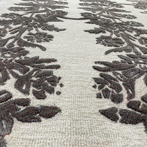 May include: A rectangular area rug featuring a cream-coloured base with a raised, dark brown floral pattern. The design consists of two vertical columns of symmetrical, textured botanical motifs that run the length of the rug surface.