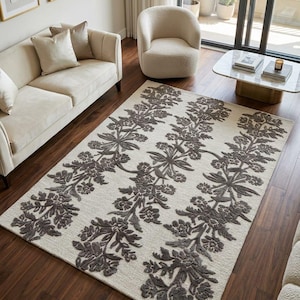 May include: A rectangular area rug featuring a cream-coloured base with three vertical columns of dark grey, raised floral and leaf patterns. The rug has a textured pile and includes a circular tag in the corner that reads Handmade in Turkey.